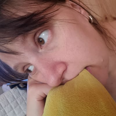 A close-up, high-angle shot focuses on the face of a person, primarily the left side, with brown hair. The subject's left eye is wide open and looking to the left, revealing a brown iris. Their nose is visible, with the right side partially obscured by a yellow, textured cloth, possibly a towel or blanket, held near their mouth. The person's skin appears pale with some redness around the nose. A small, silver earring is visible in the left ear. The background is blurred, suggesting an indoor setting with soft lighting. The image is cropped tightly to the face and the yellow cloth, with only a hint of a quilted surface visible in the lower left. There is no information in the image that suggests a location in Dunnae-myeon, South Korea.