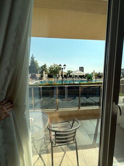 A hand is pulling open a light-colored curtain on the left side of the frame, revealing a view of a resort-like area with a swimming pool and lounge chairs. The view is from a balcony or patio, indicated by a glass railing with metallic supports and a tiled floor. In the foreground of the balcony, there is a round glass-topped table with a metallic base and a metallic chair.
Beyond the railing, the swimming pool is a prominent feature, filled with blue water. Around the pool, white umbrellas provide shade for lounge chairs. A green inflatable slide is visible near one end of the pool. Trees and some buildings, including a tall, dark building in the distance, are visible beyond the pool area under a clear blue sky. The overall scene suggests a sunny day at a recreational facility, likely a hotel or resort in Hisarya, Bulgaria.