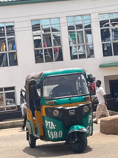 In Bukuru, Nigeria, a three-wheeled auto-rickshaw, painted green and yellow, is parked on a paved road in front of a white building. The auto-rickshaw has "PTA-6988" written on its side. A man in a white t-shirt and tan pants is walking away from the auto-rickshaw to the right. In the background, the building has multiple windows with reflections of the sky and possibly interior objects. Some of the windows show what appear to be clothing or banners hanging inside. To the right of the building, a dark doorway is visible. A large, rough stone is situated on the right side of the frame.