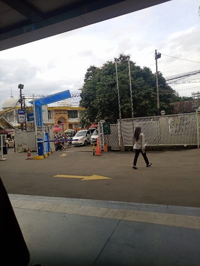 The image captures an outdoor urban scene in Bogor, Indonesia, in the vicinity of Balai Kota Bogor (Bogor City Hall). The view is from beneath a dark, overhead structure, looking out onto a paved asphalt surface under an overcast sky.
In the immediate foreground, a blue-grey paved area with a light grey stripe transitions to an asphalt road. A large yellow directional arrow is painted on the asphalt, pointing left.
In the midground, a female pedestrian is walking from right to left across the asphalt. She wears a light grey long-sleeved top, dark pants, and a grey hijab, holding a dark, rectangular object in her right hand. To the left, a controlled entry point features blue and yellow automated barriers. Two orange traffic cones are positioned near these barriers. Behind the barriers, a queue of motorbikes is visible, followed by at least two white vans, one bearing blue markings. A tall, white, slatted fence extends across the right midground, behind which stand three vertical silver poles and a large, dense green tree.
The background is dominated by the yellow-facade building of Balai Kota Bogor, identifiable by its arched entrance and visible signage, positioned centrally behind the vehicle queue. To its left, a white-domed structure, characteristic of a mosque, is partially visible, alongside various market stalls indicated by colorful umbrellas. ATM signs for "BSI" and "mandiri" are attached to a pole near the dome. Utility poles and overhead wiring are present against the cloudy sky on the right side of the frame.