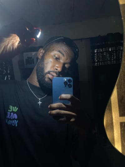 The image, taken in Lokoja, Nigeria, shows a man in a dimly lit room. He is holding a smartphone and a small flashlight, which illuminates his face and hand. He is wearing a black t-shirt with colorful text that appears to read "WE ARE YOUR". A silver chain with a crucifix pendant is visible around his neck. The background is dark and appears to be a wall with some reflective surfaces. The man is looking at his phone, suggesting he is taking a selfie or recording a video.