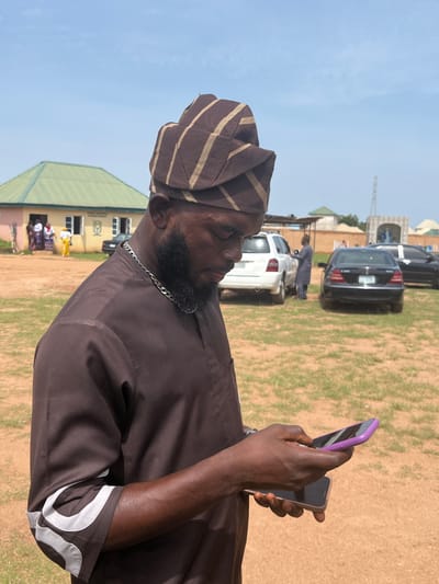 A man wearing a brown and gold striped cap and a brown tunic stands in an outdoor area with dry, sandy ground and sparse grass, looking down at two mobile phones in his hands. He has a dark beard and is wearing a silver chain around his neck. In the background, a yellow building with a green roof is visible, with a few people standing near the entrance. Several cars are parked in the background, including a white SUV, a black Mercedes-Benz sedan, and another black car. The sky is clear and blue, with a few wispy clouds. The location appears to be a public space, possibly a community center or a gathering place, in Jos, Nigeria.