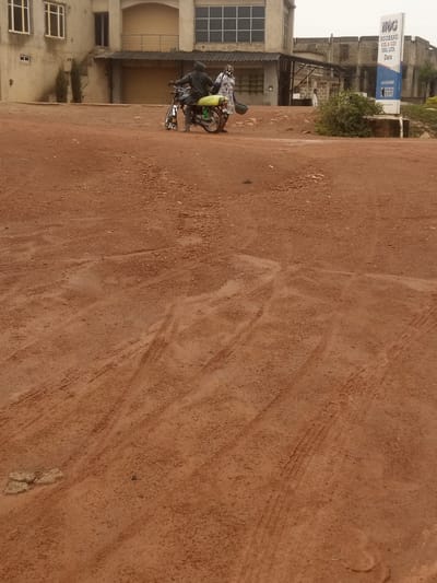 The image depicts a reddish-brown, dirt road in front of a partially constructed building. A motorcycle with two individuals on it is parked on the road, facing left. The driver is wearing a dark jacket and a hooded garment, and a yellow bag is strapped to the back of the motorcycle. The passenger is wearing a patterned dress and carrying a blue bucket. To the right of the motorcycle, a sign for "NGOBANG OIL & GAS NIG. LTD. Dura" is visible. The building behind the motorcycle appears to be a commercial or office structure with multiple windows and a balcony. The environment suggests an outdoor, possibly semi-rural or developing urban area. The ground is uneven and shows tire tracks, indicating recent vehicle activity. The sky is overcast, contributing to a muted lighting condition. This scene is located in the city of Kwang, Nigeria.