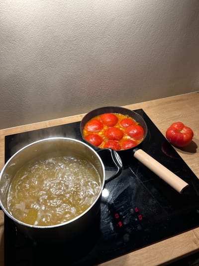 This image depicts a kitchen counter scene in the Archanes Municipal Unit, Greece, featuring active food preparation. On the left, a silver-colored metal pot with a black handle is positioned on a black ceramic induction stovetop, containing vigorously boiling, yellowish-brown liquid from which steam rises. To its right, a black frying pan on the same stovetop holds approximately eight whole or halved red tomatoes simmering in a light-colored liquid. Further to the right, a single large, red, irregularly shaped tomato rests directly on the light-colored wooden countertop. A light-colored, cylindrical object, resembling a kitchen tool handle, lies on the stovetop between the boiling pot and the loose tomato. The induction stovetop displays red illuminated numbers, including "0," "9," and "6," indicating active heating elements. The stovetop surface exhibits visible smudges. The background consists of a textured, light beige wall above the wooden countertop. No individuals are visible.