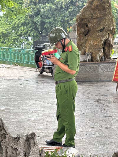 An individual, appearing to be a police officer in a green uniform and helmet, stands on a paved walkway, looking down at a smartphone. The officer is wearing a dark belt and black shoes. To the left of the officer is a red basket filled with what appear to be yellow fruits, resting on the seat of a parked black scooter. The scooter is parked near a green metal fence. In the background, a large, weathered rock formation stands prominently, with carved decorative elements at its base. Lush green trees are visible behind the fence, and the ground is wet and appears to be paved with patterned tiles. A red sign with white text is partially visible on the right side of the frame. The overall scene suggests an outdoor setting, possibly a tourist area or a public space within Hanoi, Vietnam, on a day with overcast skies or recent rain.