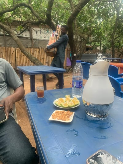 A Nigerian man, likely in his early twenties, is captured carrying a stack of boxed items. He is seen from the waist up, wearing a dark blue tunic-like garment. The man is walking past a bamboo fence and under a canopy of trees. The background reveals a partially constructed building, suggesting the location might be an outdoor market or a less developed area.
In the foreground, a blue plastic table is set with refreshments. A clear glass contains a amber-colored liquid, possibly juice or iced tea. Beside it, a white plate holds several slices of lime or lemon, and another small white dish contains a pile of roasted peanuts or cashews. A large, decorative white and silver thermos jug sits on the table, along with a clear plastic bottle of water. A person's arm, clad in a grey short-sleeved shirt and dark denim jeans, is visible on the left side of the frame, holding a smartphone. The ground appears to be sandy or dirt. The overall setting is informal and appears to be an outdoor seating area, possibly at a local eatery or relaxation spot. The scene is likely set in Abuja, Nigeria, given the context provided.