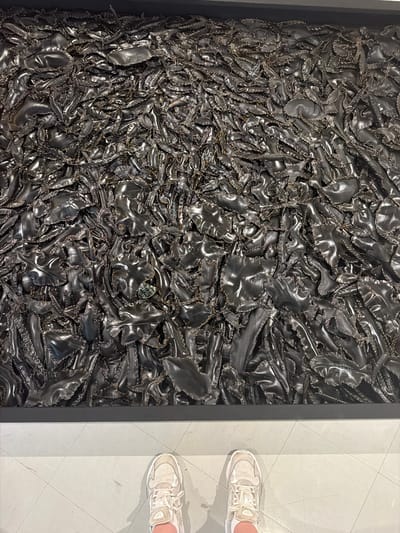 An overhead, full-frame shot shows a massive art installation made of black, shiny, textured material, possibly recycled rubber, pieced together with visible stitching. The material is arranged in a dense, organic, and somewhat chaotic pattern, filling the entire frame from edge to edge, with some pieces appearing to overlap and fold over one another. The surface has a metallic sheen, reflecting ambient light and creating highlights and shadows that add depth and texture. In the foreground, at the bottom of the image, a person's feet are visible, wearing a pair of light-colored athletic shoes. They are standing on a light gray tiled floor, with faint lines marking the tile grout. The art installation is framed by a dark, horizontal border at its base. The scene appears to be an indoor exhibition space, possibly in a gallery or museum. The city of Bangkok, Thailand, is not directly depicted in the image, nor is there any information in the visual data to confirm its location.
