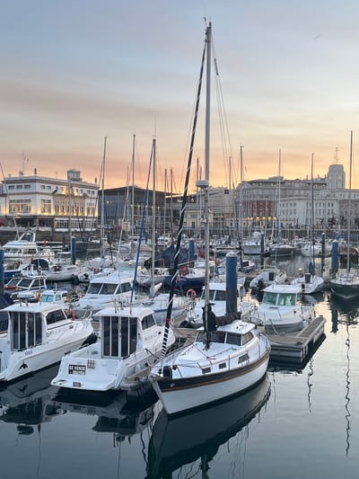 This is a photograph of a marina in A Coruña, Spain, taken at dusk. The sky is a soft gradient of blue and orange, indicative of sunset. Numerous sailboats and motorboats are docked, their masts reaching towards the sky. Some of the boats are white, while others have darker hulls. Reflections of the boats and their masts are visible on the calm water of the harbor. In the background, several multi-story buildings are visible, some with lit windows, suggesting urban development surrounding the marina. One building appears to be a distinct white structure with arched windows. The overall atmosphere is tranquil and serene, characteristic of a marina at the end of the day.