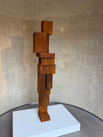 This image shows a top-down view of an abstract sculpture made of rusted metal cubes and rectangular prisms. The sculpture is arranged in a linear fashion, with the elements gradually increasing in size towards the right end, where it rests on a white pedestal. The sculpture is set against a backdrop of a textured concrete floor. To the right of the sculpture, the floor transitions to a darker tiled surface, bordered by a curved metal edge. The lighting suggests an indoor environment, possibly a museum or gallery. The overall scene is clean and minimalist, highlighting the geometric form and material of the artwork. There is no visible text in the image. The location is identified as The Hague, Netherlands.