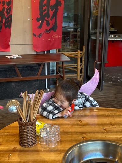 A child sits at a wooden table playing with a small water pistol. The child has a high ponytail and wears a checkered shirt, with what appears to be a purple blanket or cape behind them. On the table are a metal hot pot, several small glasses, and a woven basket containing what look like chopsticks and a ladle. Red banners with black Chinese characters hang on the wall nearby. The setting appears to be a restaurant. The city and country have been specified as Jiangjin, China.