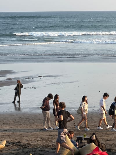 A group of people are walking on a beach in North Kuta, Indonesia. The ocean is in the background with gentle waves. The sky is clear and the sun is setting. The beach is wet sand with some rocks. The people are dressed in casual clothing. Some are walking in pairs or alone. One person is sitting on the sand with a drink.