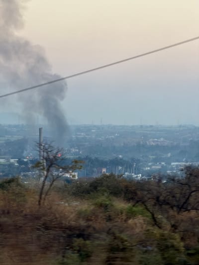 An industrial cityscape shrouded in haze features a tall smokestack emitting dark smoke. The sky is overcast, and a transmission line cuts diagonally across the view. Residential buildings are visible amidst trees, but visibility is poor due to the air quality. The foreground contains foliage, blurring the image. The overall impression is one of pollution and environmental impact.