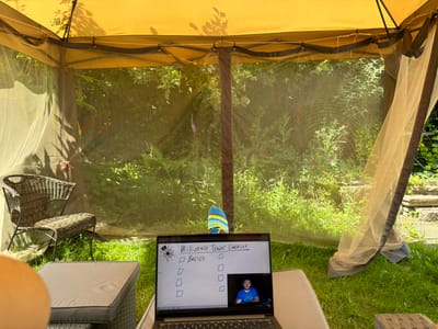 The image shows a person's feet, wearing a striped sock, visible from the knees down, resting on a cushioned outdoor seat within a screened gazebo. The gazebo has a yellow roof and sheer netting on the sides, allowing a view of lush green foliage and grass. In the foreground, a laptop is open on another cushioned seat, displaying a "Millionaire Trader Checklist" with checkboxes and a video call window showing a man with a beard wearing a blue t-shirt. To the left of the laptop, a woven armchair is placed on the grass. The setting appears to be a backyard in a suburban or semi-rural environment, bathed in natural sunlight. There is no specific information to indicate the location is in Leeds, United Kingdom.