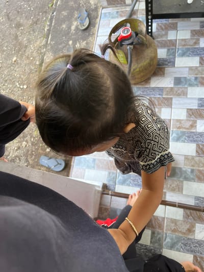 A young child with dark hair styled in two pigtails is captured from a high-angle perspective, looking down at the ground. The child is wearing a black and white patterned shirt and black pants, with a thin gold bracelet on their right wrist. They are standing on a tiled floor with a pattern of beige, brown, and grey squares. To the left of the child, a person wearing a dark grey garment is partially visible, their hand extended towards the child. In the background, a rusty-colored gas cylinder with a red regulator is connected to a pipe. The ground outside this area appears to be a rough, grey surface, with a single blue flip-flop visible near the edge. The scene is outdoors, with a glimpse of a railing or edge in the foreground.