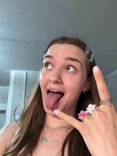 A woman with light brown hair and fair skin is posing for a selfie indoors. She is sticking her tongue out with wide eyes and a playful expression. She is wearing blue hair clips in her hair, a gold ring with a pink gemstone, and a ring with pink and white beaded flowers. She also has on a delicate necklace with a heart-shaped pendant. The background appears to be a bathroom with a gray wall and white tiled surfaces. The location in Perm, Russia cannot be verified.