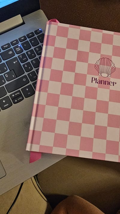 The image shows a close-up, overhead view of a pink and white checkered planner and a portion of a laptop keyboard. The planner is placed on top of the laptop. The planner has a shell icon and the word "Planner" printed on its cover. A pink ribbon bookmark is visible extending from the bottom of the planner. The laptop keyboard has various keys, including "Supr," "Num Lock," "Inicio," and number keys. A blue light is illuminated on the laptop keyboard, possibly indicating power or a status light. The background appears to be a textured surface, possibly a desk or table. The image is static and there are no people clearly visible. The location is not specified in the image.