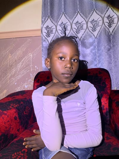 The media file shows a young Black girl lying on a red velvet chair. She is wearing a light purple long-sleeved shirt and denim shorts. Her head is resting on her arms, and her eyes are looking directly at the camera. She has a neutral expression on her face. The background consists of a patterned curtain and a wall with a splattered design. The lighting suggests an indoor setting, possibly in the evening or at night, with a spotlight creating a yellow-orange halo effect on the left side of the image. The girl's hair is styled in braids. The overall scene appears to be a casual, personal photograph. No visible text is present in the image.