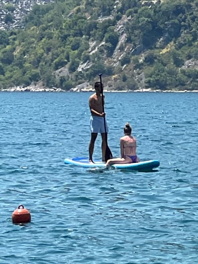 In the Bay of Kotor, near the town of Risan, Montenegro, two individuals are seen on a paddleboard. A shirtless man in light blue shorts stands at the rear of the paddleboard, holding a paddle. A woman in a bikini sits on the front of the paddleboard. The paddleboard is on a body of water with ripples. In the foreground, a red buoy floats in the water. In the background, a steep, tree-covered hillside slopes down to the water's edge. The sky is bright and appears to be a clear day.
