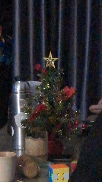This image depicts an indoor environment in Thành phố Đà Lạt, Vietnam, characterized by low light and visible digital noise.

The central object is a small artificial Christmas tree, adorned with green foliage, red decorative elements (appearing as small baubles or clusters), and gold tinsel. A gold star serves as the tree topper. The tree rests within a visible red pot.

To the left of the Christmas tree, a tall, cylindrical stainless steel object, possibly a thermos or insulated container, stands on the surface. Next to this, closer to the foreground, is a light-colored planter, appearing to be ceramic or concrete, featuring a wavy or scalloped upper rim and containing green foliage distinct from the Christmas tree.

In the bottom left corner, a partial view of a light-colored, possibly ceramic, mug is visible. In the bottom right corner, a multi-colored Rubik's Cube is present, displaying blue, yellow, and orange faces, with some white squares visible.

All objects are arranged on a flat, dark, and somewhat reflective surface, likely a table. The background consists of dark, vertically pleated or striped material, consistent with curtains or blinds. No individuals are visible, and no actions or interactions are depicted within the static scene.