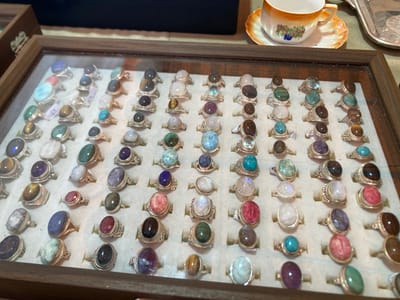 The image displays a close-up, eye-level view of a wooden tray filled with an assortment of rings, each featuring a distinct gemstone. The rings are arranged in neat rows and columns on a light-colored, velvety surface within the tray. The gemstones vary in color, shape, and size, including shades of blue, green, purple, pink, white, and brown. Some stones are smooth and cabochon-cut, while others appear faceted. The metal settings of the rings are silver-toned, with intricate detailing visible on some bands. In the background, a portion of a decorative teacup and saucer, and a metallic tray are visible, suggesting a retail or display setting. The lighting suggests an indoor environment, possibly a shop or market stall. There are no people or animals visible in the image, and no specific location in Barcelona, Spain, can be identified.