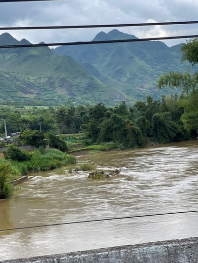 The image depicts a wide, muddy river flowing through a valley in Lùng Tám Commune, Vietnam. On the left bank, there is a small village with houses and a power pole, interspersed with lush green vegetation and what appear to be agricultural fields. The river itself has a choppy surface with visible currents and a few scattered rocks emerging from the water. The right bank is densely covered with various types of trees, including bamboo. In the background, a range of steep, verdant mountains dominates the landscape under a cloudy sky. The image is framed by two dark horizontal wires in the foreground and a textured concrete railing at the very bottom.