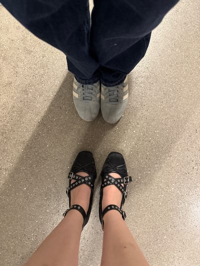 The image shows a view from above of two pairs of feet standing on a speckled light-grey surface. On the left, a pair of feet is wearing blue jeans and grey sneakers with white stripes. On the right, a pair of feet is wearing black ballet-style flats with multiple straps and eyelet details. The setting appears to be an indoor space with artificial lighting, possibly in Mexico City, Mexico.