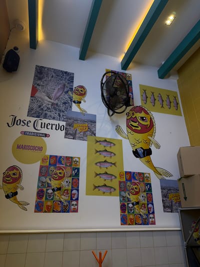 The image shows a wall in a restaurant or business in Mexico City. The wall is white with various decals and posters. Some of the notable decorations include:
* Decals of fish with Lucha Libre masks.
* Posters of fish in rows.
* A poster of Mexico City.
* A poster with the words "Jose Cuervo Tradicional Marisococho" in large print.
* A poster featuring a hand holding a fish superimposed over a city map.
A black stand-up fan is affixed to the wall. The location appears to be indoors, and possibly a casual dining establishment.