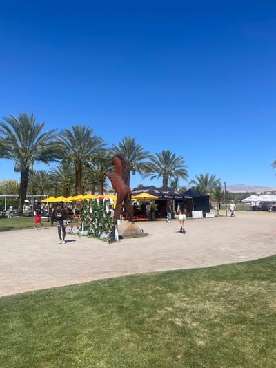 A partly sunny outdoor scene in Thermal, United States, with a clear blue sky and numerous tall palm trees is depicted. The scene includes a bronze-colored horse sculpture, vendor stands, and various decorative elements, including yellow umbrellas and floral arrangements. People are present, either sitting or standing near the vendor stands. The ground comprises a brick pathway and a green grassy area. In the background, a mountain range is visible, alongside some buildings.