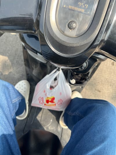 The image shows a close-up, point-of-view shot from someone riding a motorcycle. The rider's blue jeans and white Croc-style shoes are visible. A white plastic bag with red and yellow lettering is hanging from the handlebars. The motorcycle's dashboard displays a digital readout of "959". The background appears to be a street in Batang, Indonesia, with a neutral gray asphalt surface. The lighting suggests it is daytime with bright sunlight.