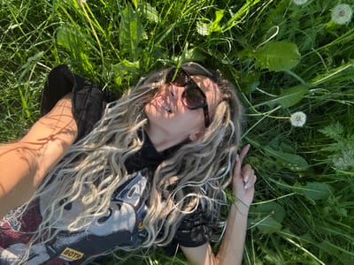 The image depicts an adult individual, appearing female, lying supine in a dense patch of sunlit green grass located in Rusanovo, Russia. The subject's head is tilted upwards, looking towards the sky. She wears dark sunglasses and has long, wavy blonde or light brown hair interwoven with lighter, possibly braided, strands, spread out on the grass. Her mouth is slightly open, revealing teeth. She is dressed in a black short-sleeved t-shirt featuring a prominent graphic design on the front, with a yellow rectangular label reading "BOTR" in black text visible on the lower left side of the shirt. Her left arm (viewer's perspective) is bent with the hand resting near her head, while her right arm (viewer's perspective) extends slightly outwards, fingers lightly curled. Her fingernails are manicured with a light, possibly glittery, polish. Distinct shadows from overhead foliage are cast across her skin and clothing. The grassy environment includes various broad-leafed plants and several white, spherical dandelion seed heads.