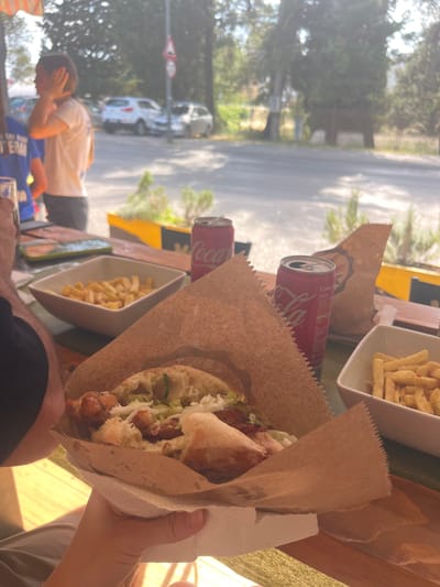 The image shows a partially eaten kebab wrapped in brown paper held in a hand. Next to the kebab are a can of Coca-Cola, a dish of fries, and another kebab wrapped in paper. In the background, there is a street scene with trees, cars, and a pedestrian crossing sign. A person is standing by the street with their head in their hand. The location is likely in Podgorica, Montenegro.