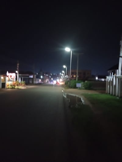 The image captures a street scene at night in Gura Top, Nigeria. A paved road dominates the foreground, reflecting the light from several street lamps that line the left side of the road. On the left side of the road, there is a brightly lit establishment with a red and white facade and outdoor seating. Further down the road on the left, additional structures with lights are visible. Across the road on the right, a building with a dark roof is partially obscured by a white fence-like structure. Beyond the buildings, there are trees and bushes. The sky is dark, indicating nighttime, and the overall atmosphere is one of a quiet, illuminated street.
