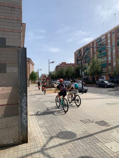 The image depicts a sunny day in Valencia, Spain, on a wide sidewalk paved with light-colored square tiles. In the foreground, three young people are riding bicycles on the sidewalk. The person closest to the viewer is wearing a black t-shirt with a graphic print and a red helmet, riding a black bicycle. To their left, another person with a blue backpack is riding a teal bicycle. Further down the sidewalk, a third person is riding a bicycle with an orange frame. All three are moving in the same direction, away from the viewer.
The sidewalk is bordered by a street with parked cars and moving vehicles. Several multi-story brick buildings with green awnings line the opposite side of the street, characteristic of urban architecture in Valencia. Some trees and streetlights are also visible. On the left side of the image, a large metal fence with a diamond pattern dominates the foreground, casting a patterned shadow on the sidewalk. Graffiti is visible on the metal support pole of the fence. The sky is a clear blue with a few wispy clouds. The scene suggests a casual urban activity on a pleasant day.