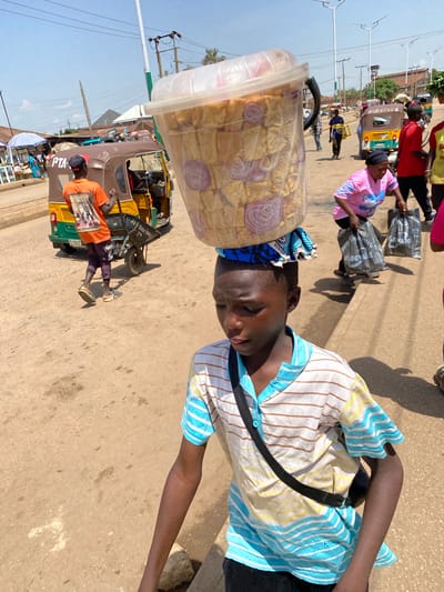 In Bukuru, Nigeria, a young Black boy walks forward, carrying a large, clear plastic container filled with food items, balanced on his head. The container is covered with a blue and white patterned cloth. He wears a striped short-sleeved shirt and dark pants, with a black strap across his chest.
The environment is a sun-drenched, dusty urban street. In the background, a colorful auto-rickshaw, a common mode of transport, is visible. Other people are present, including a woman in a pink shirt carrying shopping bags and a person in an orange shirt pushing a cart. The scene depicts a bustling market or street scene.
