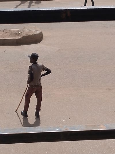 A young male stands in profile on a dirt road, facing away from the camera and looking to the left. He is wearing a dark baseball cap, a light-colored t-shirt, and reddish-brown trousers. A long, thin stick is held vertically in his right hand, and his left hand rests on his hip. The ground is a dusty, light brown surface, possibly a road or pathway. In the upper left of the frame, a raised, irregularly shaped object, appearing to be a concrete or stone curb or planter, is visible. The background consists of a blurred, sandy-colored area, suggesting an open space. The image is framed by a dark, metallic horizontal bar in the foreground, obscuring the bottom portion of the scene. The lighting suggests daylight, casting shadows on the ground. The location is specified as Bukuru, Nigeria.