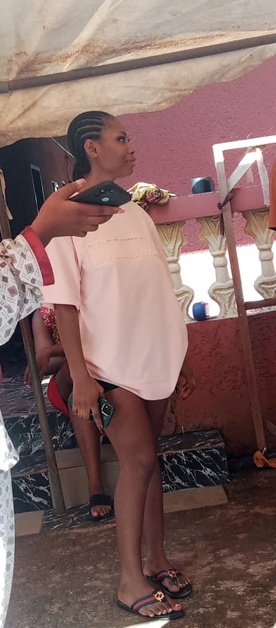 The image depicts an outdoor or semi-outdoor setting within Udi, Nigeria; a specific address or landmark is not identifiable. A young adult female with dark skin and cornrow braids stands centrally, wearing a light pink oversized t-shirt with "ESSENTIALS" embossed on the front, black shorts or underwear, and dark flip-flops featuring red, blue, green stripes, and a gold circular emblem. She holds a green-cased smartphone in her right hand and directs her gaze towards the upper right. To her left, the hand of another individual, sporting pink fingernail polish and wearing a white patterned long-sleeved garment, holds a dark smartphone with a visible triple camera array, pointing it towards the standing female, indicative of a photo or video capture. A third individual is partially visible in the mid-background, seated, wearing a red garment and black furry slides. The background features a textured maroon wall, fronted by a low, light-colored decorative balustrade with a darker red base. Above, a white, stained canvas or tarp serves as a canopy, supported by a dark horizontal bar and vertical poles. The ground is composed of concrete and sections of black-and-white marbled material. Incidental objects include a blue cylindrical item and a piece of yellow fabric resting on the balustrade.