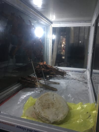 In a display case, likely a food warmer, possibly located in Gwarinpa, Nigeria, are several skewers of cooked meat, possibly suya, stacked on the left and in the middle. To the right, a stack of flatbreads (possibly roti or chapati) are partially wrapped in a yellow plastic bag. The display case appears to be in a public area, as indicated by blurred figures and a lighted interior reflecting off the glass. The case is made of metal and glass with condensation on the glass panes.