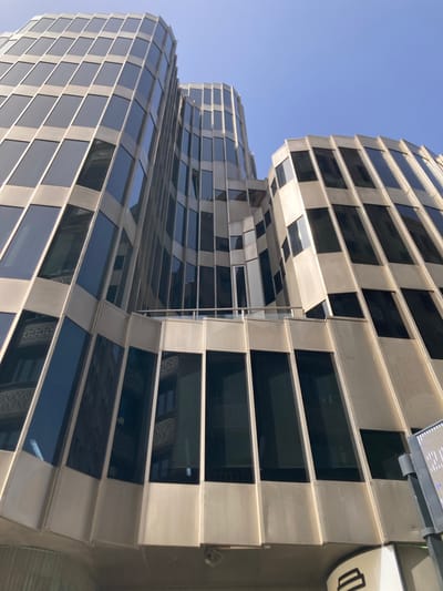 This media file shows a close-up, low-angle shot of a modern, unusually shaped building under a clear blue sky, suggesting daytime with fair weather. The building's facade is composed of alternating dark glass panels and light-colored, possibly metallic, structural elements. The glass panels reflect the surrounding environment and the sky, creating a dynamic visual effect. The building's structure appears to curve and twist, with horizontal bands of windows and structural material creating a textured, layered appearance.

In the upper right portion of the image, a section of the building shows a white, rounded sign with a black symbol that is difficult to decipher clearly, but resembles a stylized drawing. Above this sign, a rectangular fixture with lights is mounted to the building. The overall impression is of contemporary architecture with a focus on geometric patterns and reflective surfaces. There are no people visible in the frame. The image is oriented vertically, as if taken from ground level looking up. The location context is Barcelona, Spain. No specific activity or event is depicted.