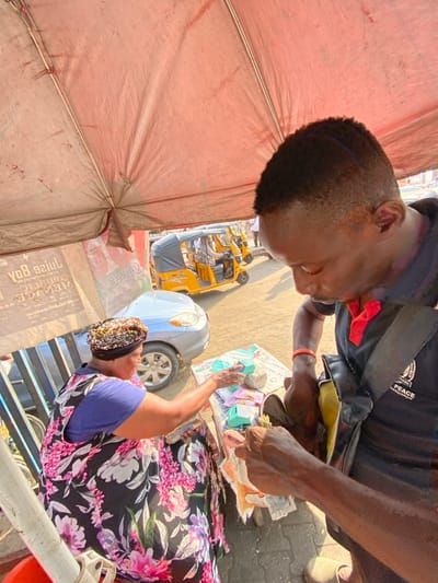 This image depicts a street vendor transaction occurring under a red canopy on a sunny day in an urban area of Lagos, Nigeria. In the foreground, a seated female vendor, wearing a multi-colored headwrap and a black and pink floral dress over a blue top, extends her right hand towards an assortment of items displayed on a surface in front of her. These items include several small, light-blue packaged boxes and other wrapped goods. Standing opposite her, a male customer, with a short haircut, wearing a dark blue polo shirt with a red collar and a black shoulder bag with a "PEACE AMBASSADOR" logo, is actively reaching into his bag with his left hand while holding some folded papers or money in his right hand.

In the mid-ground, behind the vendor, a silver sedan car is parked or slowly moving. Further back, multiple yellow Keke Napep auto-rickshaws are visible on a dirt or unpaved road, indicating active traffic. The background includes blurred commercial posters or signage. The overall scene suggests a vibrant street market environment.