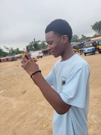 In Egbagu, Nigeria, a young man is captured in a profile view, holding a smartphone in his hands and appearing to take a photograph or video. He is wearing a light blue t-shirt with a graphic design on the front, and a black beaded bracelet adorns his left wrist. The background suggests an outdoor setting, possibly a marketplace or open area, with a dirt ground. Several auto-rickshaws, a white van, and some structures resembling small shops or stalls are visible in the distance, indicating an urban or semi-urban environment. The sky is overcast, contributing to a diffused light.