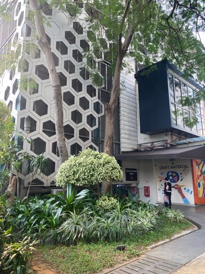 The image shows a building with a distinctive hexagonal facade, likely an art or craft supply store, given the mural on the wall that reads "ART & CRAFT MATERIAL." The building is located in North Jakarta, Indonesia. In front of the building, there is a lush garden with various green plants. A person wearing black pants and a black top with a pink bag is standing near the mural. The scene appears to be outdoors on a bright day, with trees casting shadows on the building and ground. A sign pointing downwards says "GEDUNG PARKIR MKG2," indicating a parking building. The architecture is modern with a large glass window structure on the right side of the building.