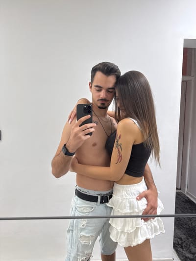 In a domestic setting, a man and a woman are taking a mirror selfie. The man, shirtless and wearing light blue distressed jeans and black underwear, holds a smartphone to capture the image. He has dark hair and a mustache. The woman, wearing a black tank top and a white ruffled skirt, has long brown hair and a tattoo on her right arm. She is embracing the man, resting her head on his shoulder and has her left arm around his neck, while her right hand is on his chest. The man's right arm is around the woman's waist, with his hand resting on her skirt. They appear to be indoors, in front of a plain white wall, with a doorway visible on the right side of the frame leading to a hallway. The overall scene suggests a private moment captured between a couple. The location is not specified in the image.