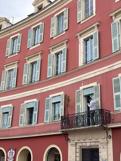 A multi-story building with a red-orange facade, consistent with architecture in Nice, France, is depicted under an overcast sky. The building features cream-colored horizontal trim lines and ornate window surrounds. Multiple windows with open, light blue-green louvered shutters are visible across several floors. On the lower floors, four windows prominently display the white capital letters "HOTEL" across their upper frames.
On the second floor from the bottom, a black wrought-iron balcony with intricate scrollwork extends from the facade. A single male individual with dark hair and a beard, wearing a white long-sleeved shirt and dark patterned pants, stands on this balcony. He is holding a smartphone with both hands and appears focused on its screen. To his left on the balcony, a small palm-like plant is visible.
Below the balcony, a dark wooden double-door entrance is marked with the number "4" to its right. To the left of the main entrance, at street level, arched window openings are present. Above one such arch, a white crest emblem featuring the text "OGC NICE" is affixed to the red wall, confirming the location as Nice. A rooftop antenna array is visible against the gray sky.