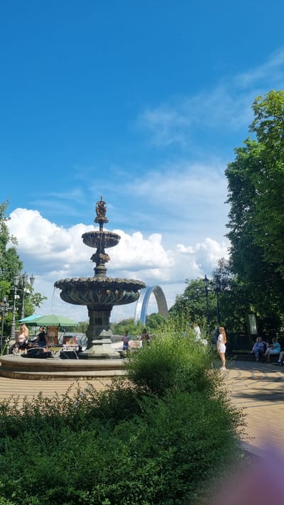 The image depicts a sunny day in a park in Kyiv, Ukraine. The scene features a multi-tiered ornate fountain in the foreground, with water spraying from its top. Behind the fountain, a large, curved, metallic structure resembling a monument or art installation is visible against a bright blue sky dotted with fluffy white clouds. Lush green trees and dense foliage frame the scene, with a paved walkway winding through the park. Several people are present, some sitting on benches, others walking, and one woman in white is standing near the fountain. The presence of people suggests a leisurely outdoor activity. The overall atmosphere appears to be peaceful and pleasant, indicative of a warm day suitable for outdoor enjoyment. The time of day appears to be midday or early afternoon due to the bright sunlight and clear sky. No visible text is present in the image.