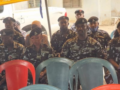 A group of seven people, all wearing camouflage uniforms and black berets, are seated in a row of blue plastic chairs under a white canopy. The person at the far left end of the row is a man with a black beret and a name tag that reads "M.S. IKEFILA". The person next to him is a woman with a name tag that reads "HFP". The person next to her is a man with a name tag that reads "J. PAUL". The man next to him is wearing a face mask. The person at the far right end of the row is obscured by a red plastic chair. The people are in a location in Maiduguri, Nigeria, near the coordinates 11.867, 13.123. The scene is captured from a low angle, and the background is blurred.