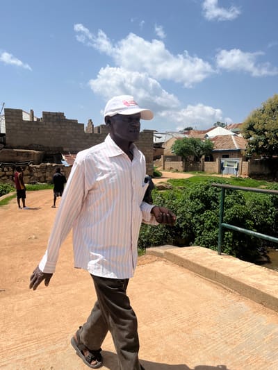 A man wearing a white baseball cap and a striped long-sleeved shirt walks on a concrete bridge in Jos, Nigeria. He is wearing dark brown corduroy pants and sandals. The man is looking to his right. In the background, there are unfinished brick buildings, trees, and houses with corrugated metal roofs. Two young men are visible in the distance on a dirt path. The sky is blue with scattered white clouds. A green metal railing runs along the side of the bridge.
