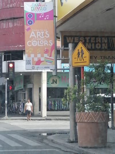 A medium shot captured from a street-level perspective shows a street scene in Davao City, Philippines. A man wearing a light-colored polo shirt and dark shorts is walking across a pedestrian lane. He is looking towards the camera. In the background, a large banner with "DUAW TA SA ARTS AND COLORS" is visible, along with a traffic light showing a red signal. Further back, shop fronts with clothing displays can be seen. A yellow pedestrian crossing sign is mounted on a pole. A large potted plant with patterned texture is situated on the right side of the frame. The overall scene depicts an urban environment with a mix of commercial activity and traffic infrastructure.