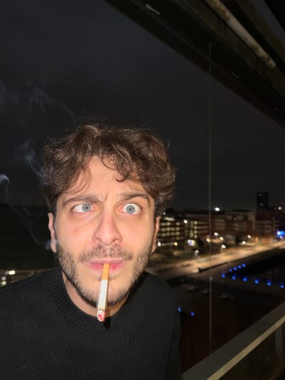 A man with curly brown hair and light eyes holds a lit cigarette in his mouth. He wears a black sweater. Smoke rises from the cigarette. The background shows a nighttime city scene with illuminated buildings and streets, suggesting a high vantage point. The image is rotated, positioning the man's head on the left.