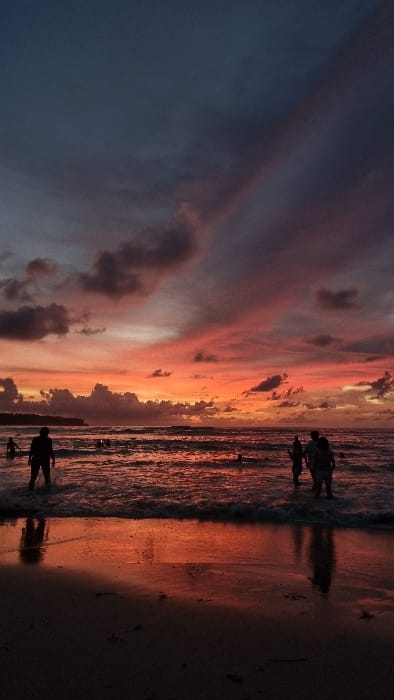 The image captures a coastal scene at dusk or sunset in Pecatu, Indonesia. The sky displays a prominent gradient, shifting from dark blue in the upper segment to vivid orange and pink at the horizon and mid-sky, interspersed with dark, scattered clouds. A distinct diagonal band of darker cloud materializes from the upper right. The foreground consists of wet sand, reflecting the reddish-orange hues of the sky and displaying subtle ripples from receding waves. The ocean water is dark with visible breaking waves near the shore, also reflecting the warm sky colors.
Approximately eight to ten human figures are present, rendered in silhouette. In the left foreground, one individual stands waist-deep in the shallow water, facing away from the viewer, with a discernible reflection on the wet sand. Towards the left and center mid-ground, multiple additional figures are partially submerged in the water, some appearing to be wading or swimming. On the right mid-ground, a group of three individuals stands together in the shallow water, facing generally left. Their lower bodies are submerged, and their reflections are visible on the wet sand. A dark landmass or cliff is silhouetted against the bright horizon on the left side of the frame.