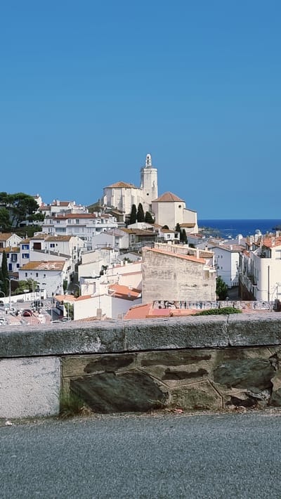 This is a vertical, wide-angle, eye-level photograph taken from a slightly elevated position, overlooking a coastal town in Cadaqués, Spain. The image is dominated by a clear, bright blue sky, suggesting a sunny day. In the foreground, a textured stone wall, composed of large, irregular blocks of grey and darker stone, runs along the right side of the frame. The wall shows signs of age and is bordered by a coarser, speckled grey surface.
The main subject is the town itself, a dense collection of white-washed buildings with terracotta-tiled roofs that cascade down a hillside towards the sea. The architecture is characterized by its Mediterranean style, with narrow streets and a charming, clustered appearance. A prominent white church with a distinctive bell tower stands out amongst the buildings, its spire reaching towards the sky. The sea, a deep blue, is visible in the distance, with a few boats dotting its surface. Some greenery, including trees, is interspersed among the houses, adding touches of color to the scene.
The image appears to be taken during the daytime, likely mid-morning or afternoon, given the bright, consistent sunlight and the absence of long shadows. The weather is clearly fair and warm. The scene conveys a sense of tranquility and picturesque beauty, typical of a Mediterranean coastal town. There is no visible text in the image.